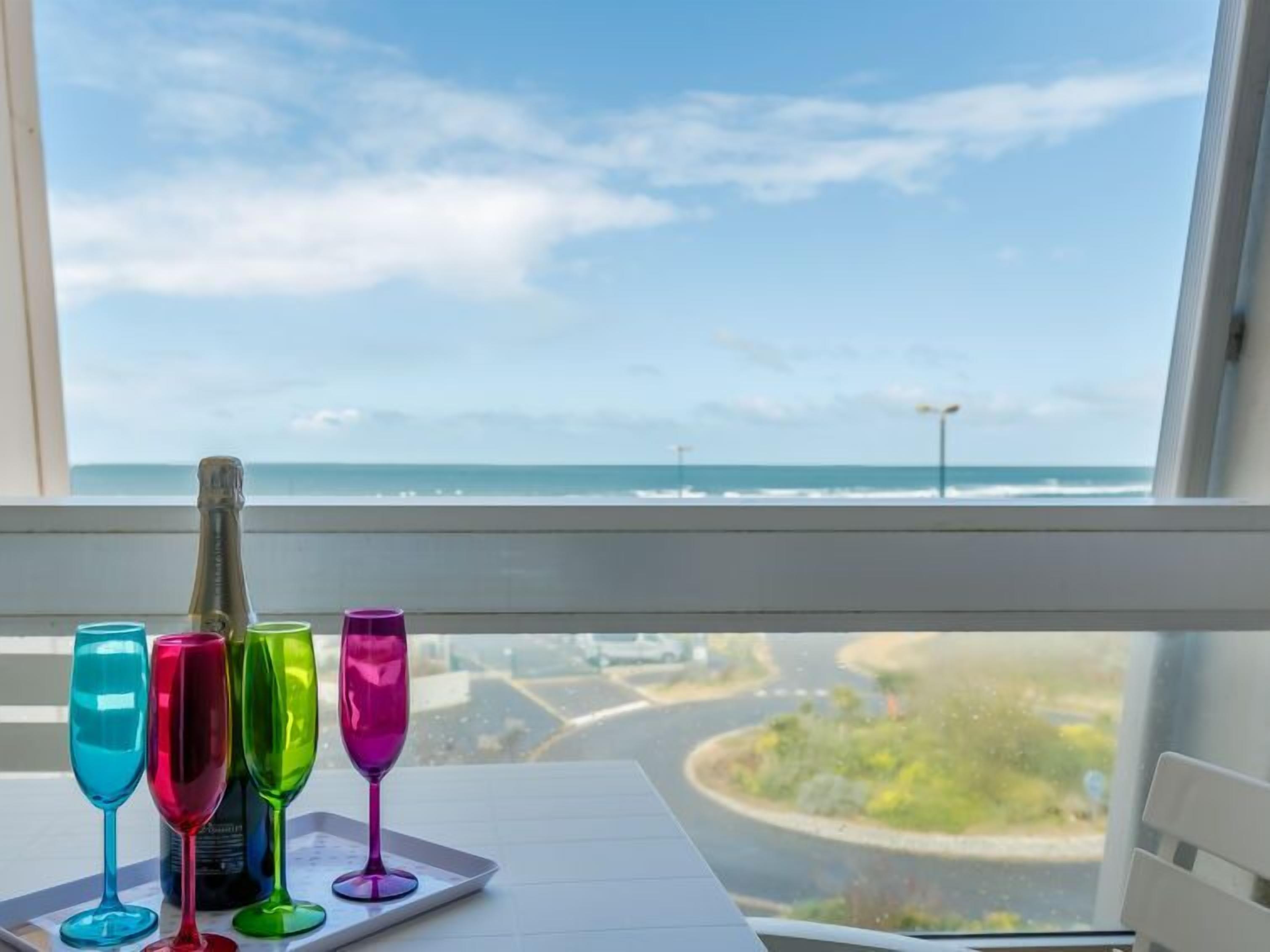 A table with colorful wine glasses and a bottle, viewed from a window overlooking a coastal road and ocean.