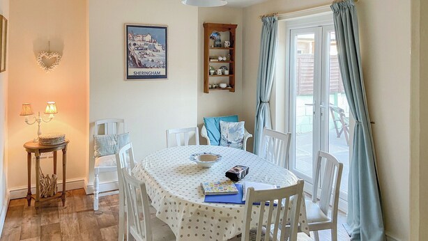A dining area with a round table covered in a polka dot tablecloth, surrounded by white chairs. There is a wall-mounted shelf with decorative items, a framed picture, and a hanging light fixture. - 3 bedroom accommodation in Sheringham (Sheringham)