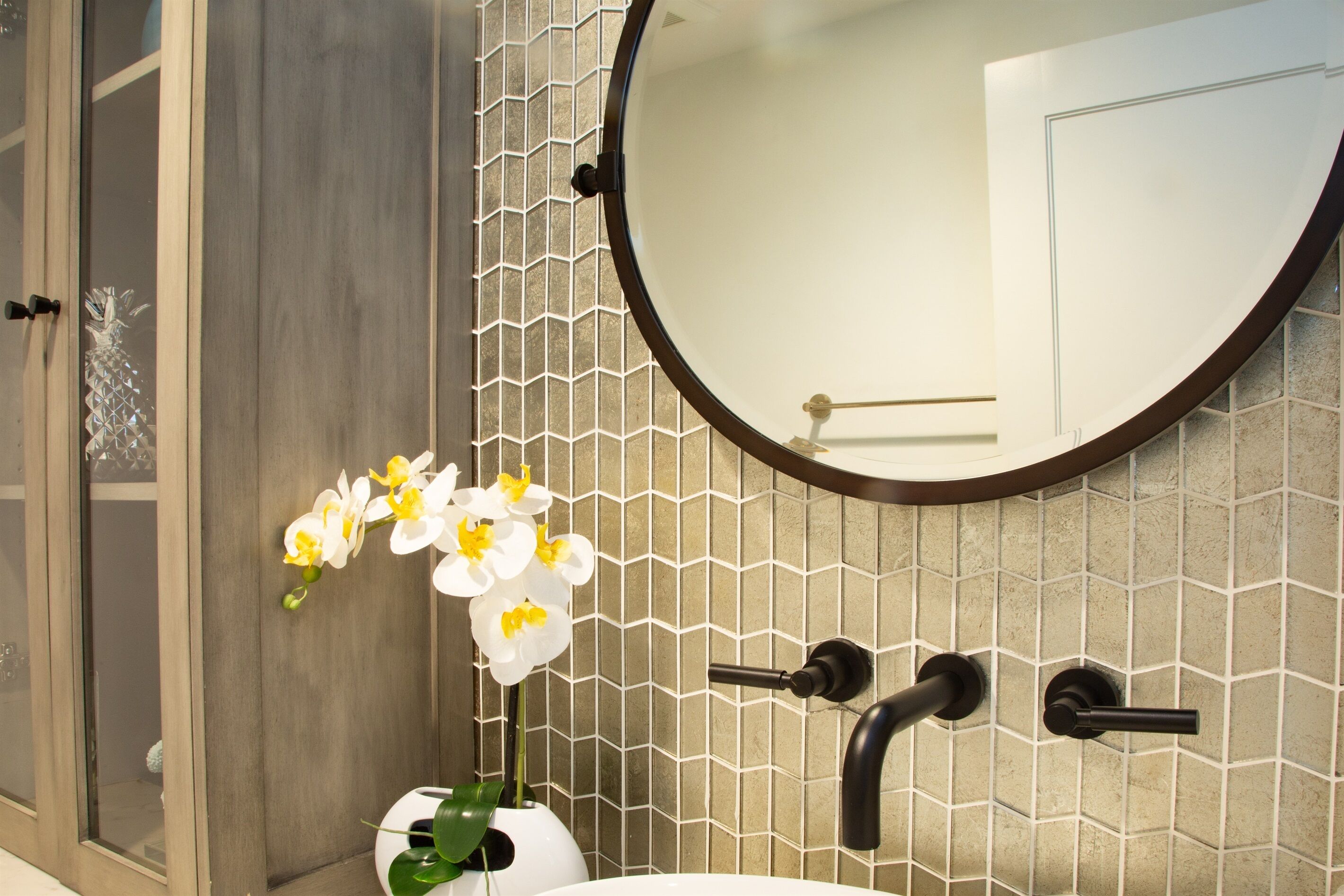A bathroom with a wooden door, a round mirror, a sink with two faucets, and a tiled wall with a geometric pattern.