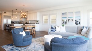 Een moderne woonkamer met een grijze bank, blauwe fauteuils en een witte salontafel. Achtergrond is een keuken met witte kasten en hanglampen.