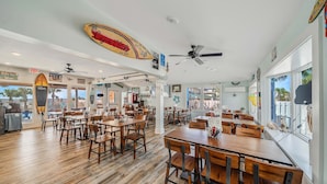 Un restaurant spacieux avec des tables et des chaises en bois, des ventilateurs de plafond et une vue sur la plage grâce à de grandes fenêtres.