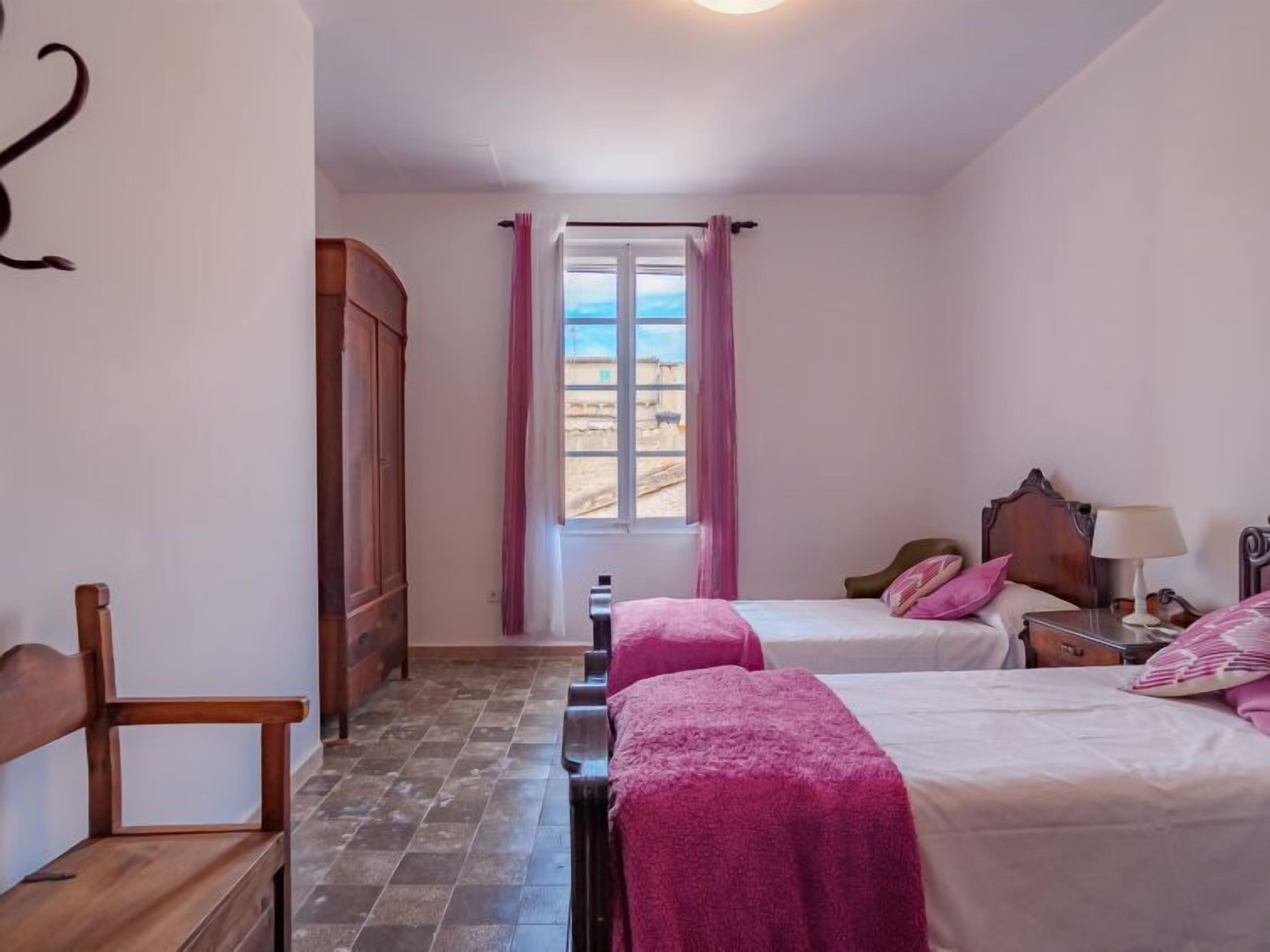 A bedroom with two beds, a wooden chair, a wardrobe, and a window with a view of the landscape.