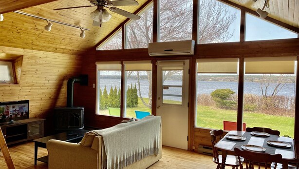 A cozy cabin with a wooden interior, a sofa, a dining table, and a large window overlooking a lake. - Cabin on the beach. (Miramichi)
