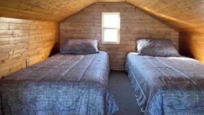 A small, cozy attic bedroom with wooden walls and a sloped ceiling. There are two single beds with matching bedding. A small window provides natural light. - Cabin on the beach. (Miramichi)