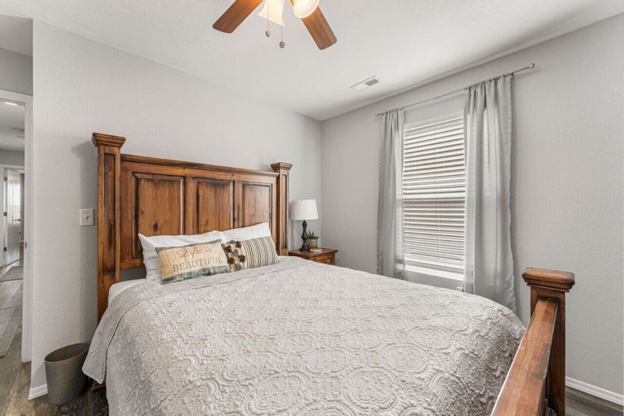 A bedroom with a wooden headboard, a bed with a patterned bedspread, a nightstand with a lamp, and a window with blinds.