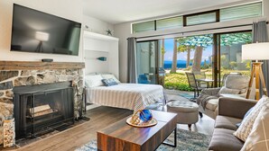 A modern living room with a stone fireplace, a large flat-screen TV, a bed with a view, and a balcony with a view of the ocean.