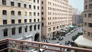Une vue sur une ville avec de nombreux immeubles, une rue animée où circulent des voitures et des piétons, et une perspective depuis un balcon doté d’une balustrade.