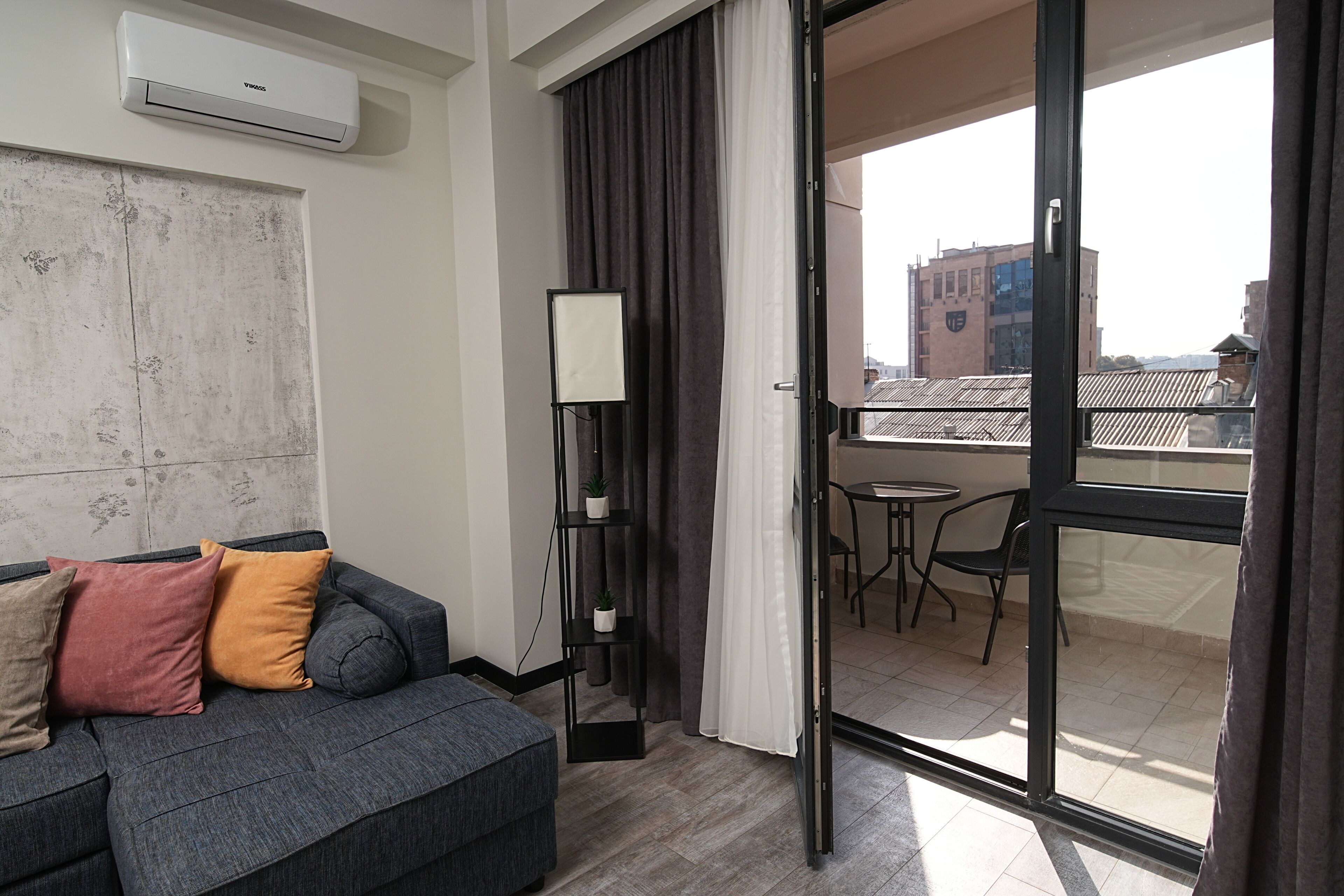 Una sala de estar moderna con un sofá gris, una mesita, una lámpara de pie y un ventanal corredero que da a un balcón con vistas a la ciudad.