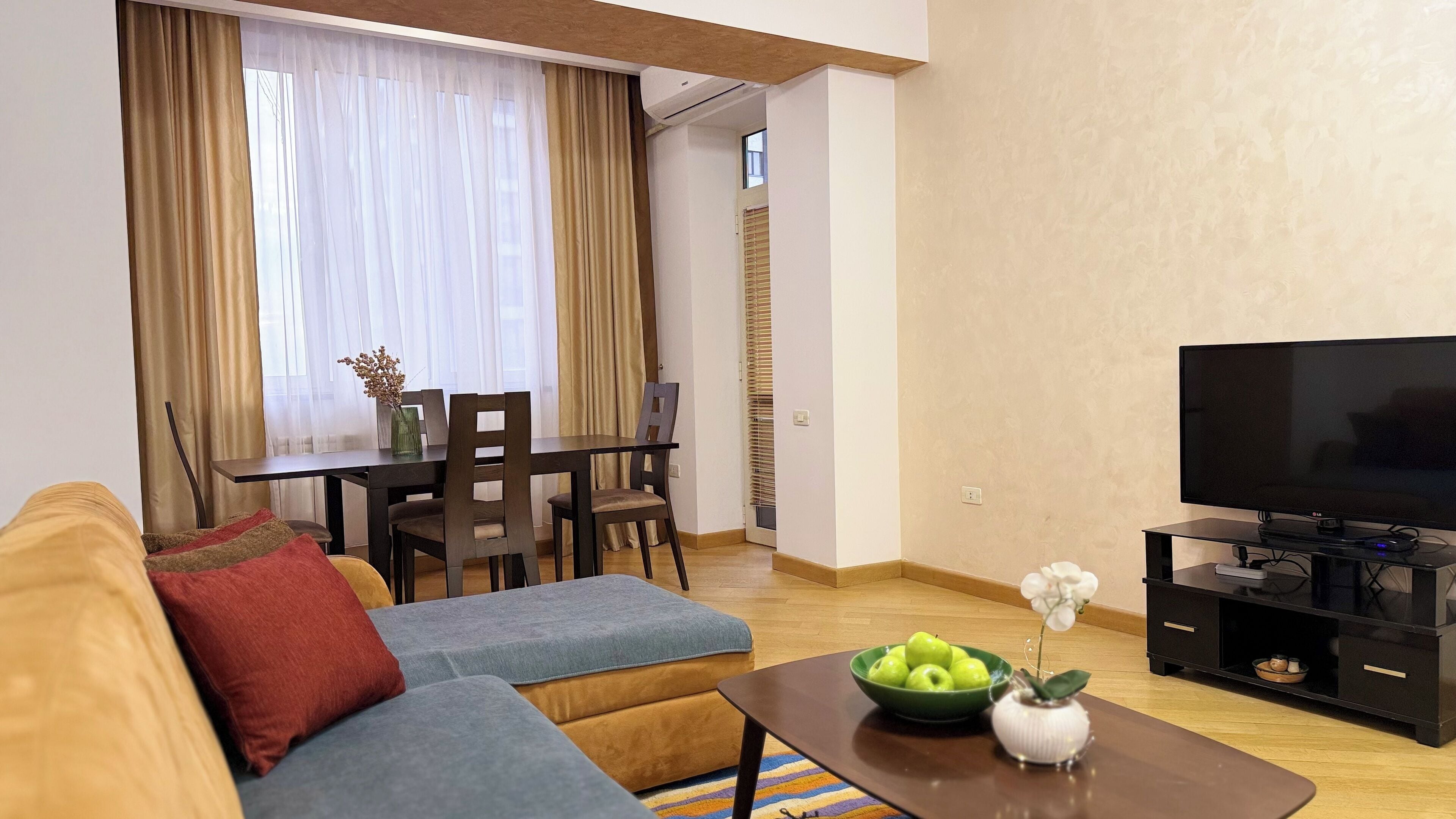 Een woonkamer met een bank, een salontafel met een schaal appels en een vaas, een televisie op een standaard, en een eethoek met een tafel en stoelen.