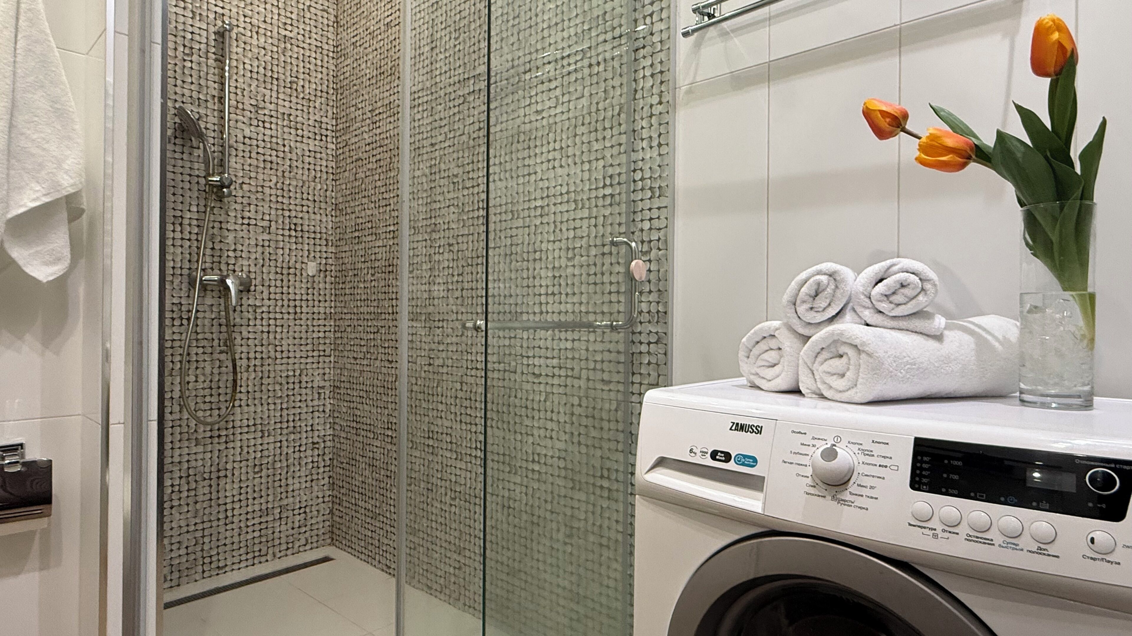 A bathroom with a glass shower enclosure, a washing machine, and a towel rack with rolled towels and a vase of tulips.