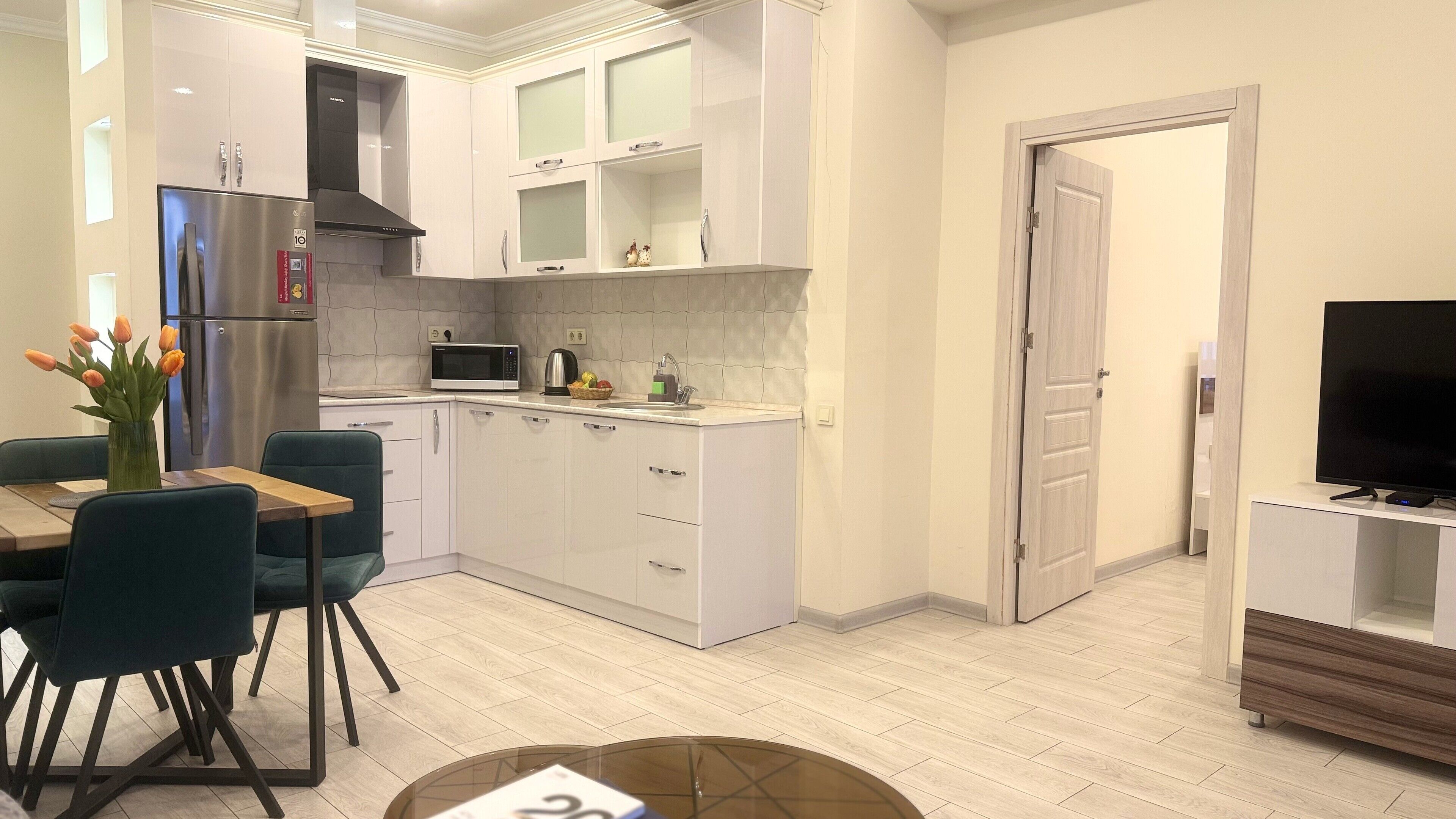 Una cocina moderna con armarios blancos, un refrigerador de acero inoxidable, un microondas y un fregadero. Hay una zona de comedor con una mesa y sillas, y un televisor en la esquina.