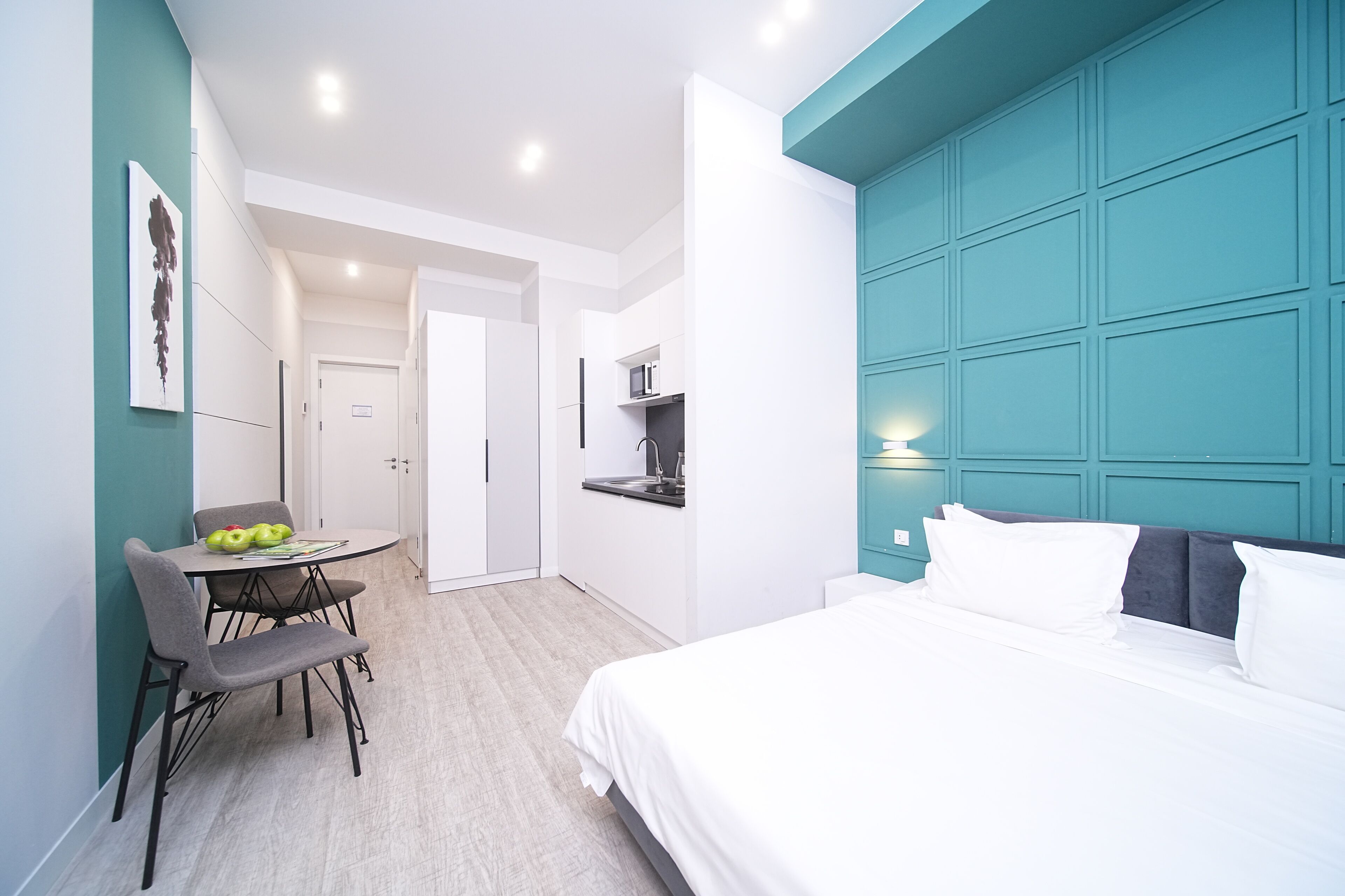 A modern hotel room with a bed, a dining table with chairs, a kitchen area, and a teal accent wall.