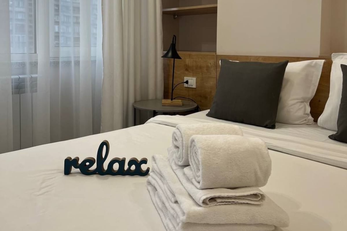 A hotel room with a bed, a bedside table, a lamp, a wall-mounted shelf, and a window with curtains.