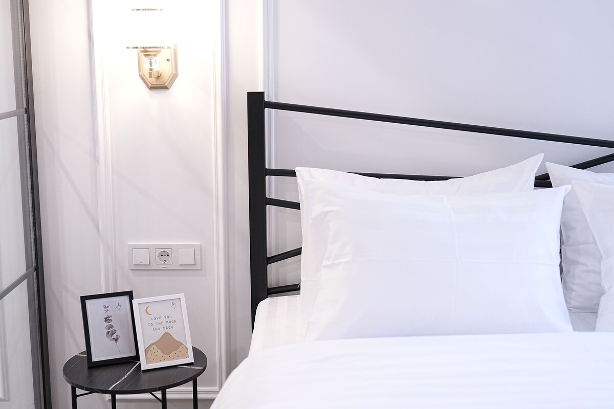 A neatly made bed with white bedding, a black metal headboard, and a small bedside table with a framed picture and a wall-mounted light.