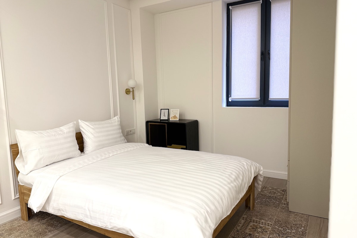 A bedroom with a bed, a nightstand, a wall-mounted lamp, and a window with blinds.