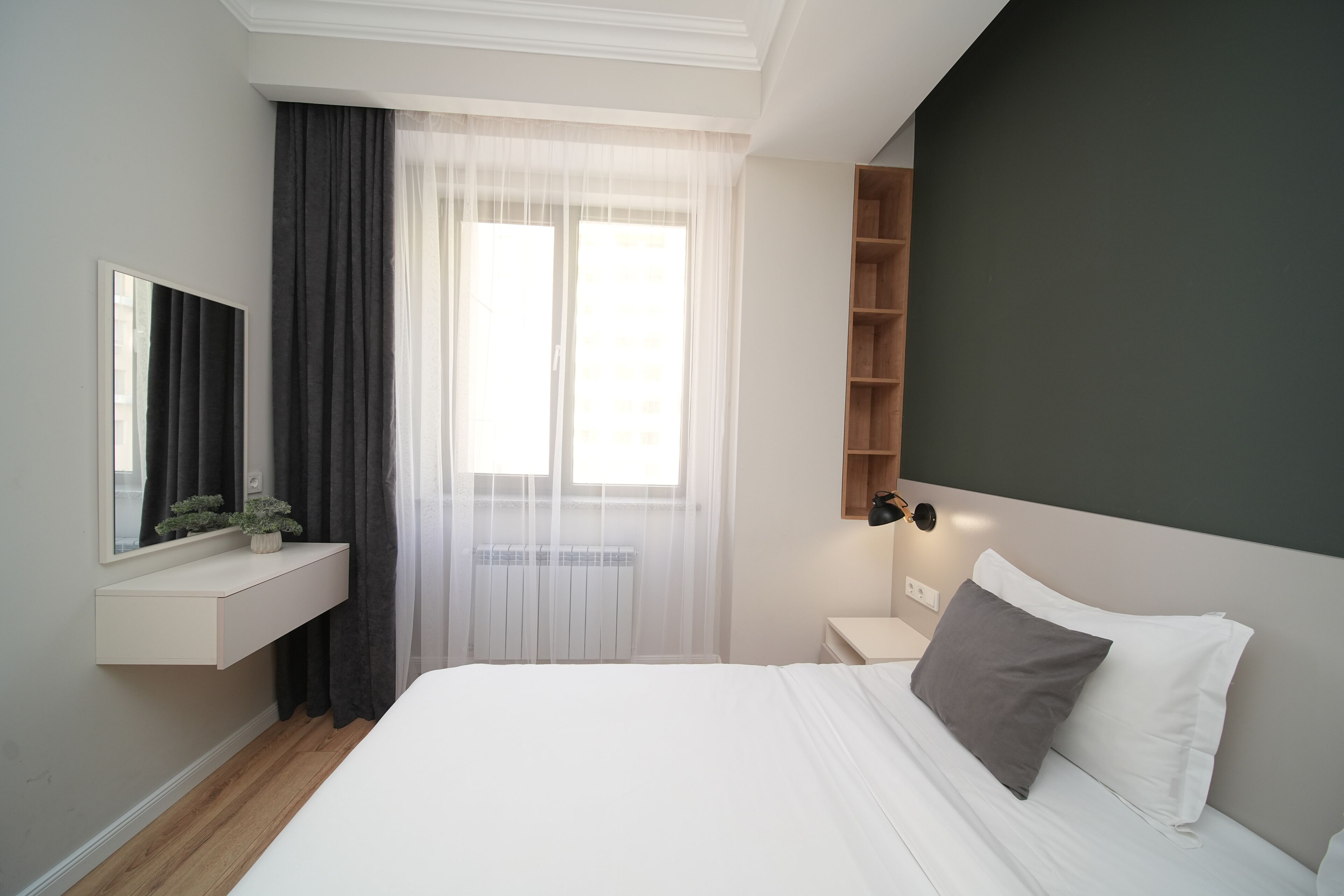 A modern bedroom with a large bed, a bedside table, a built-in shelf, a mirror, and sheer curtains.