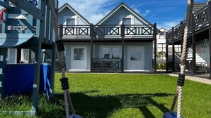 A playground with swings and a slide, a grassy area, and a building with a balcony in the background. - Holiday Home in Pobierowo near Beach (Rewal)
