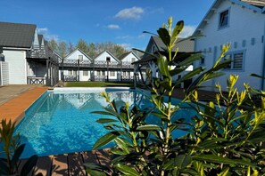 A swimming pool with a wooden deck, surrounded by a white building with a balcony.