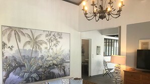 Uma sala de estar com um grande mural tropical, um sofá de vime, uma mesa de centro de madeira, um lustre e uma televisão sobre um móvel de madeira.