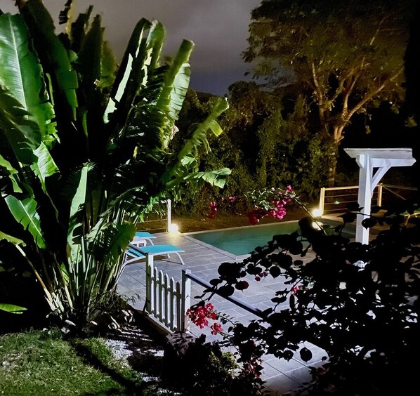 A swimming pool area with a pergola, surrounded by lush greenery and flowering plants.