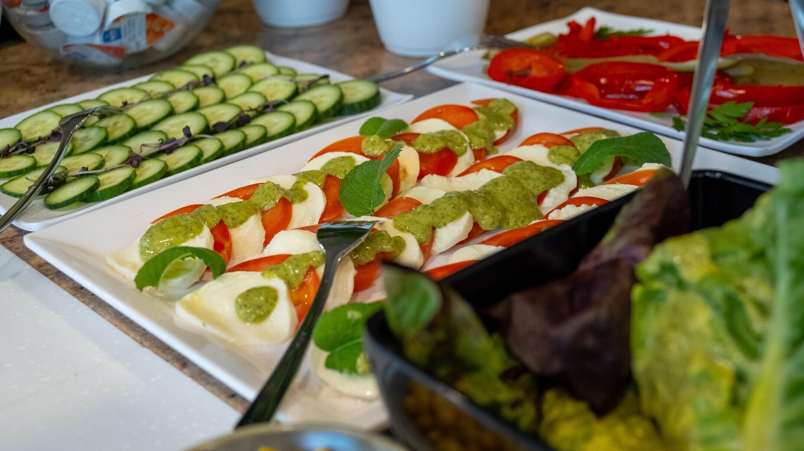 Un bufé con diversas opciones de comida, incluyendo pepinos en rodajas, tomates cherry, queso mozzarella y una salsa verde.