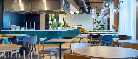 A modern restaurant interior with round tables and chairs, a central counter with a stainless steel range hood, and pendant lighting.