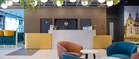 Un espace d’accueil d’hôtel au design moderne, comprenant un comptoir d’accueil, un coin salon avec des fauteuils et une grande fresque murale.