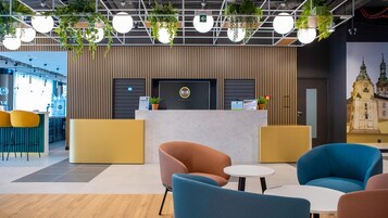 Un área de recepción de hotel con diseño moderno, que incluye un mostrador de recepción, zona de asientos con sillas y un mural grande en la pared.