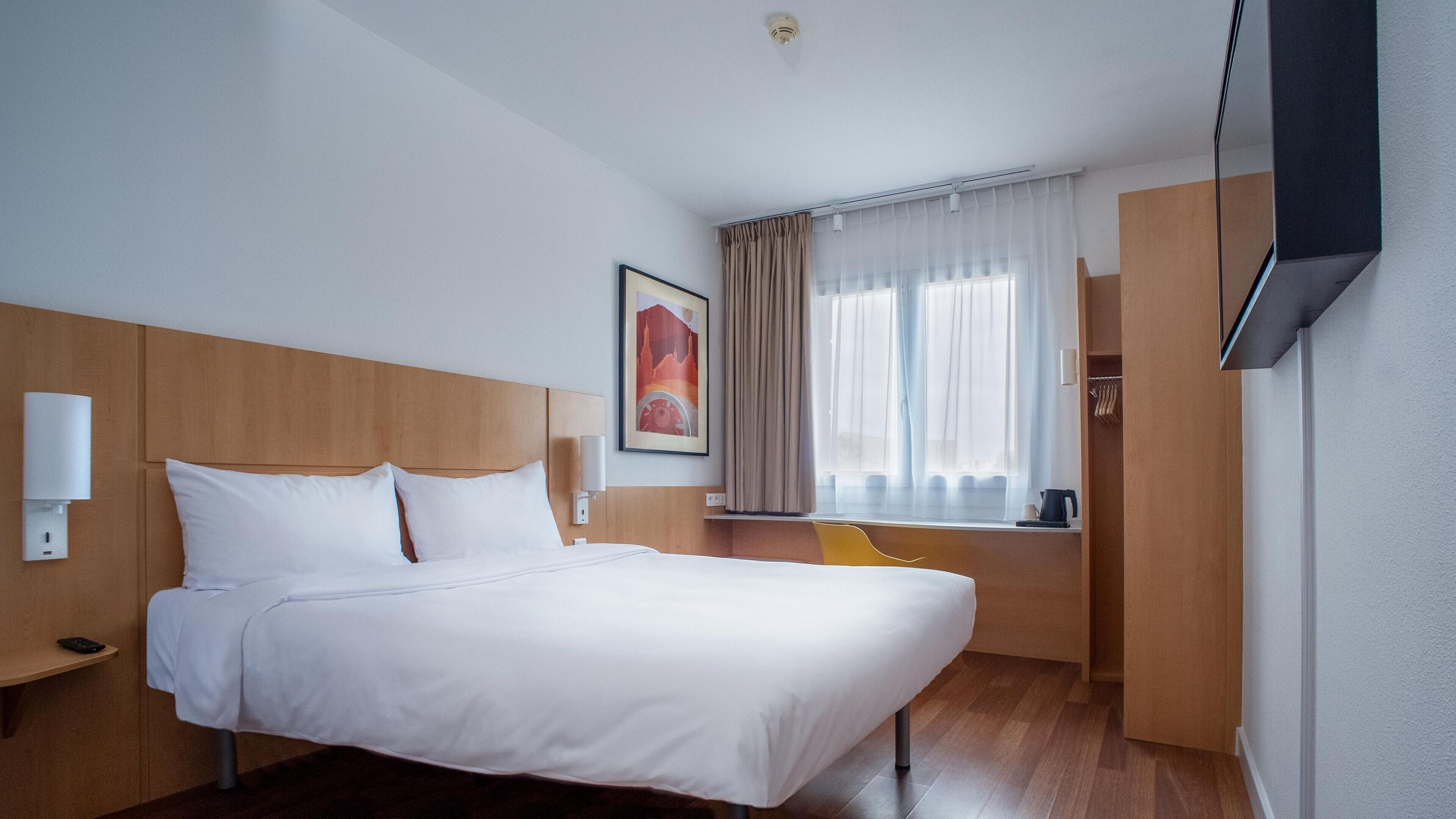 A hotel room with a bed, a bedside table, a chair, a window with curtains, a painting on the wall, and a television mounted on the wall.