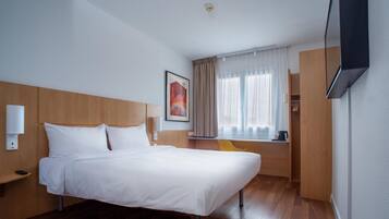 A hotel room with a bed, a bedside table, a chair, a window with curtains, a painting on the wall, and a television mounted on the wall.