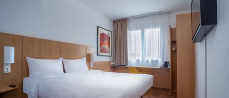 Une chambre d’hôtel comprenant un lit, une table de chevet, une chaise, une fenêtre avec des rideaux, un tableau accroché au mur et un téléviseur fixé au mur.