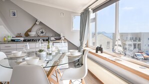 A modern kitchen with a glass dining table, white chairs, and a view of the sea through large windows. - The Penthouse @ No 5 (Lyme Regis)