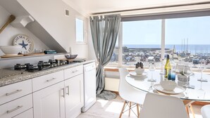 A modern kitchen with a view of the sea, a dining area with a table set for two, and a large window with curtains. - The Penthouse @ No 5 (Lyme Regis)