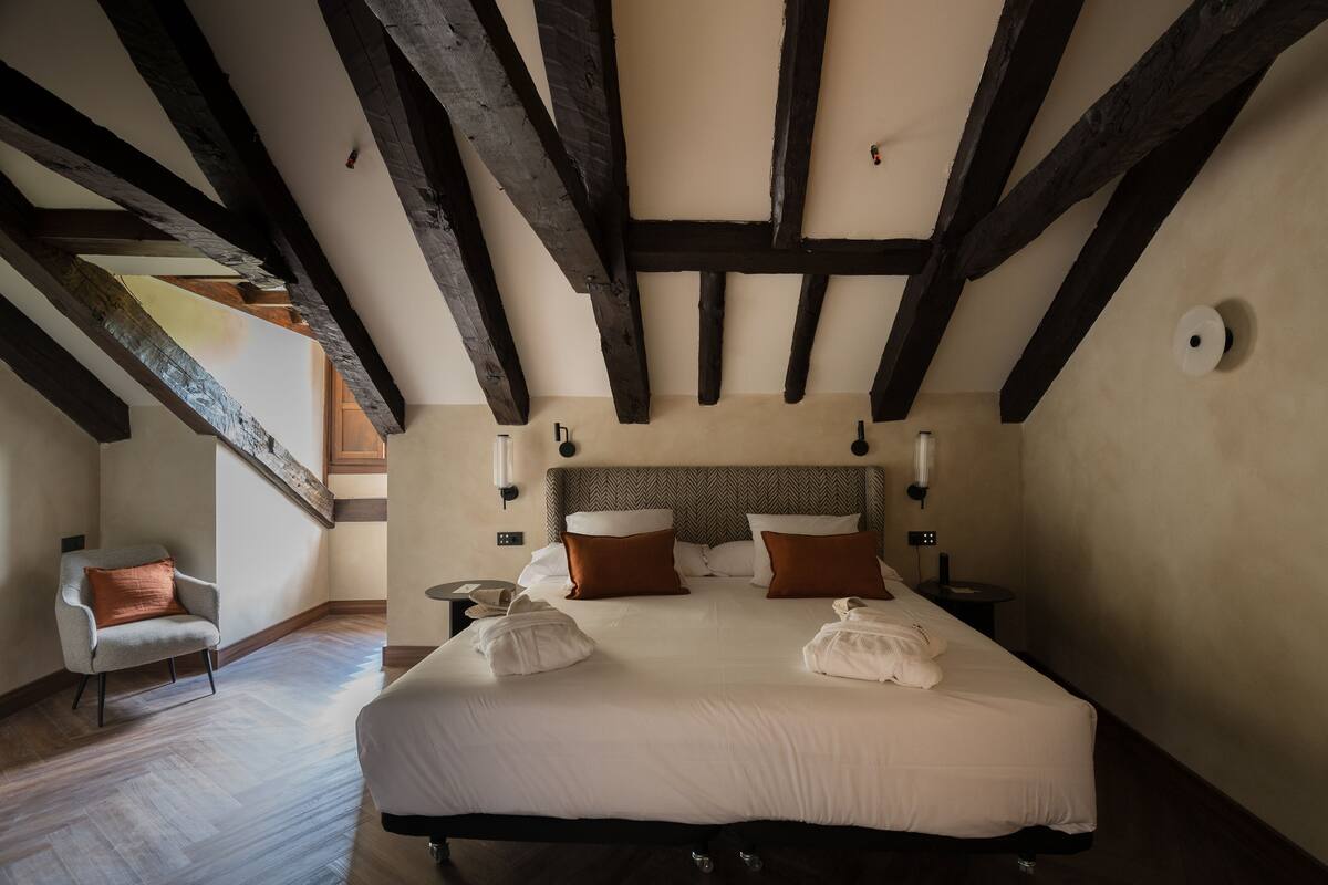 Een slaapkamer met een groot bed, houten balken, een stoel, nachtkastjes en een lamp.