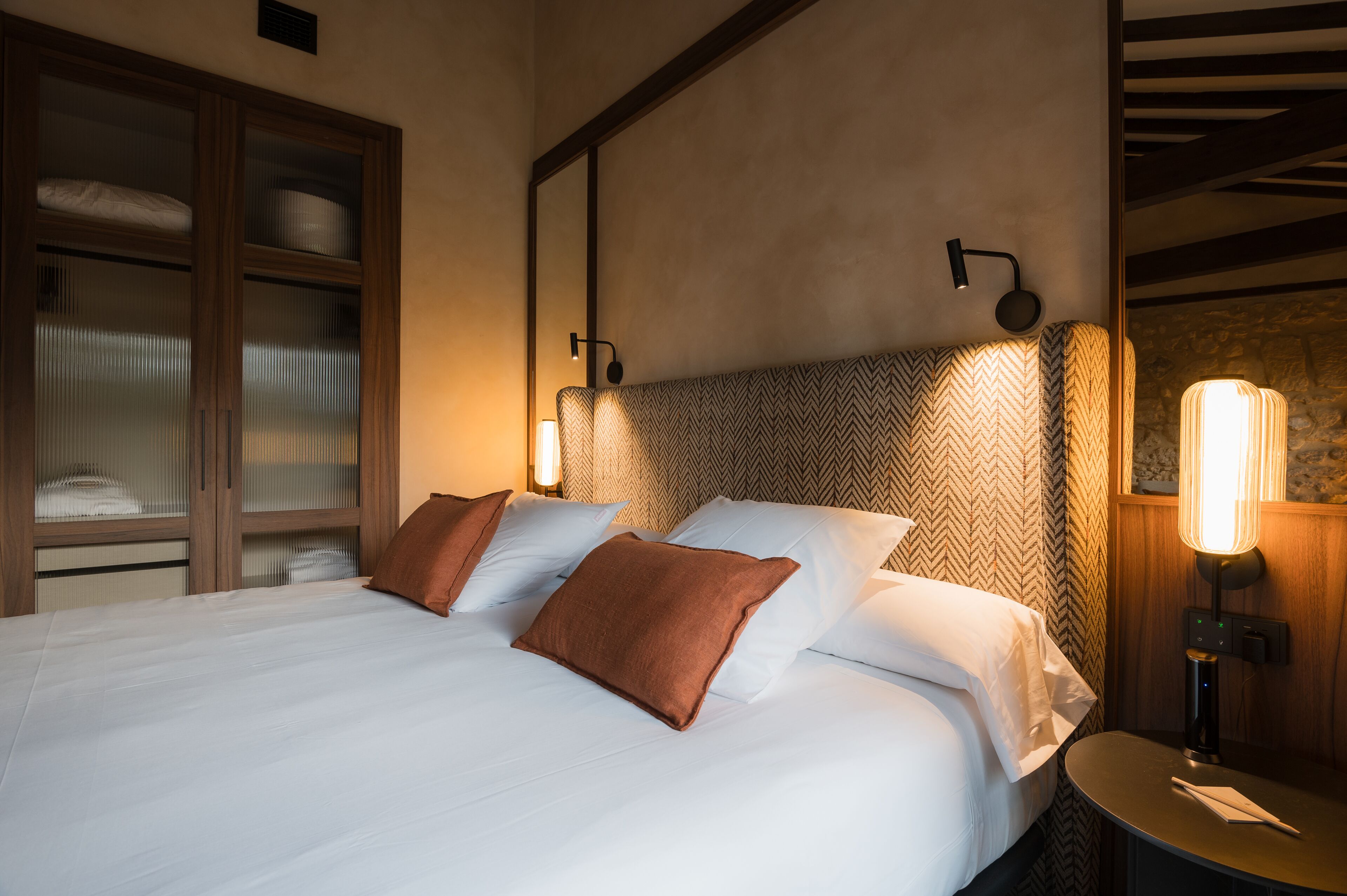 Una cama bien hecha con sábanas blancas y almohadas marrones, un armario de madera con puertas de vidrio y una lámpara fijada a la pared.