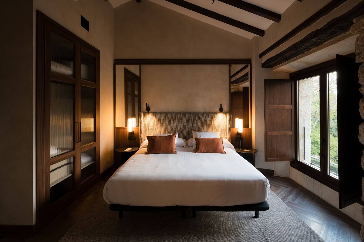Een groot bed met twee kussens, een houten hoofdbord, bedlampjes en uitzicht door een raam.