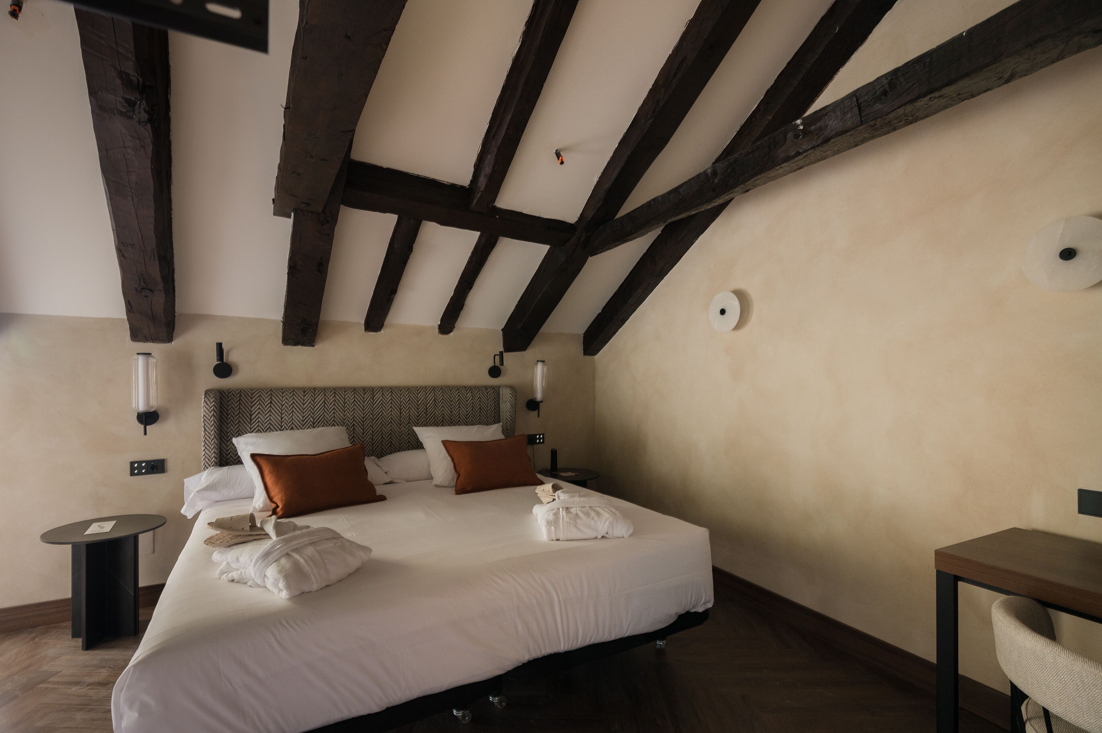 A hotel room with a large bed, wooden beams on the ceiling, a round wall clock, and a small table with a chair.