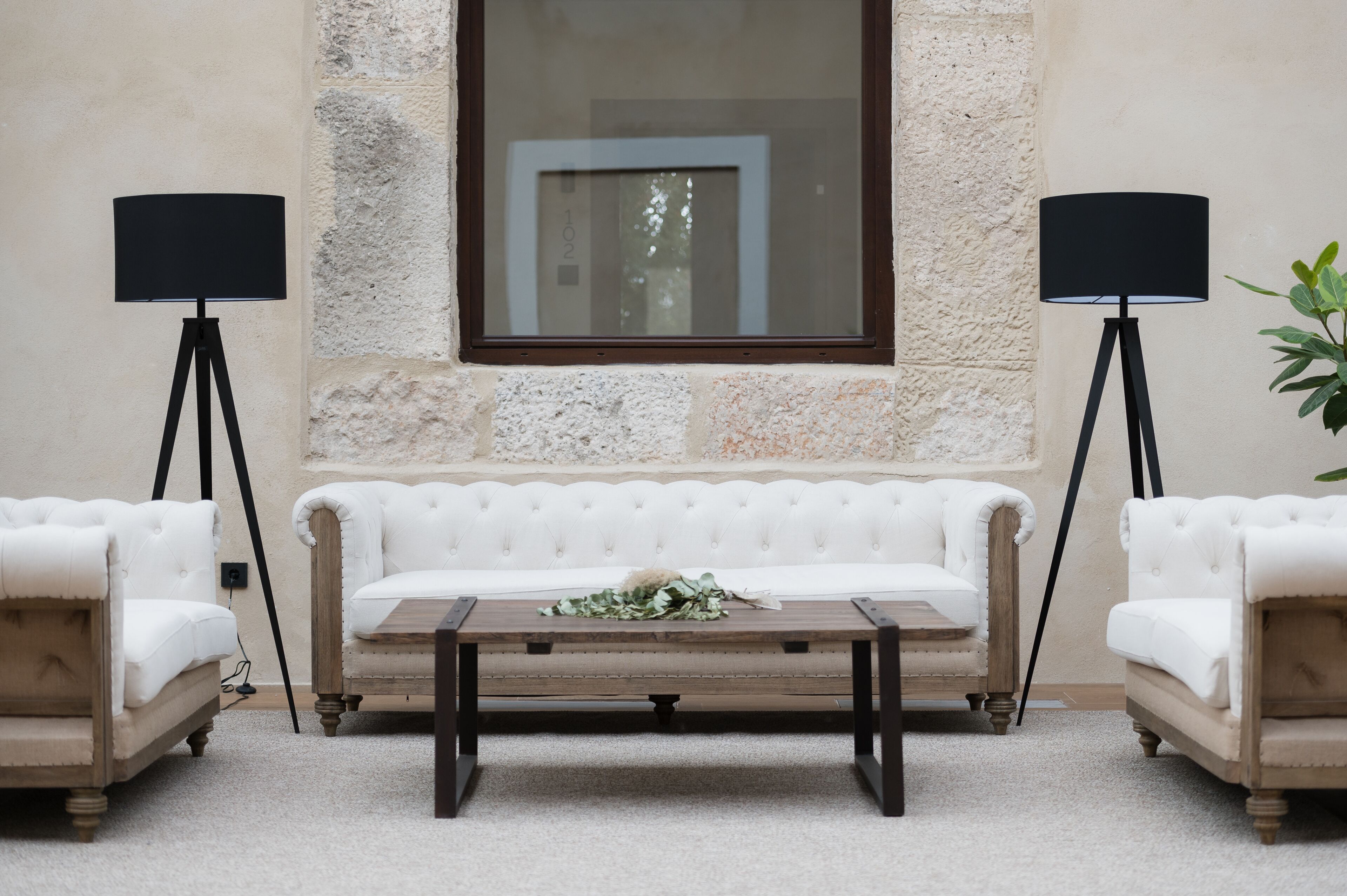 Un sofá blanco con cabeceros, acompañado por una mesa de centro de madera, dos lámparas de pie a los lados, todo frente a una pared de piedra con textura y una imagen enmarcada en la parte superior.