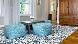Une pièce avec un tapis à motifs, une table sur laquelle est posé un vase de fleurs et des poufs bleus à motifs.