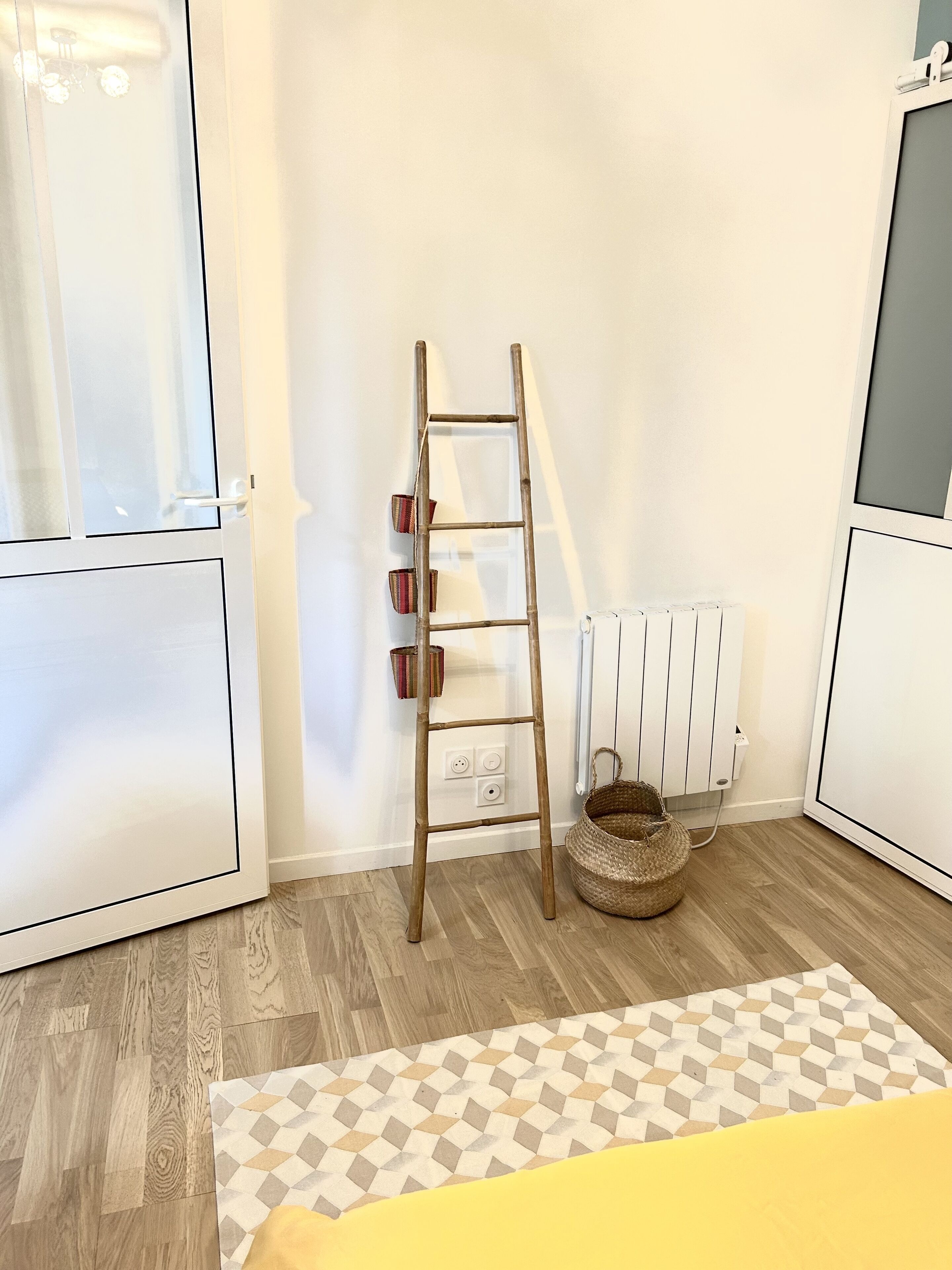 Une pièce avec une échelle en bois appuyée contre un mur blanc, un radiateur, un panier en osier et un tapis à motifs sur un plancher en bois.