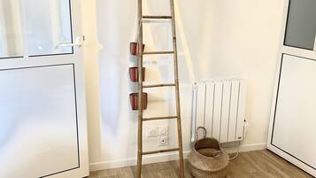 Une pièce avec une échelle en bois appuyée contre un mur blanc, un radiateur, un panier en osier et un tapis à motifs sur un plancher en bois.