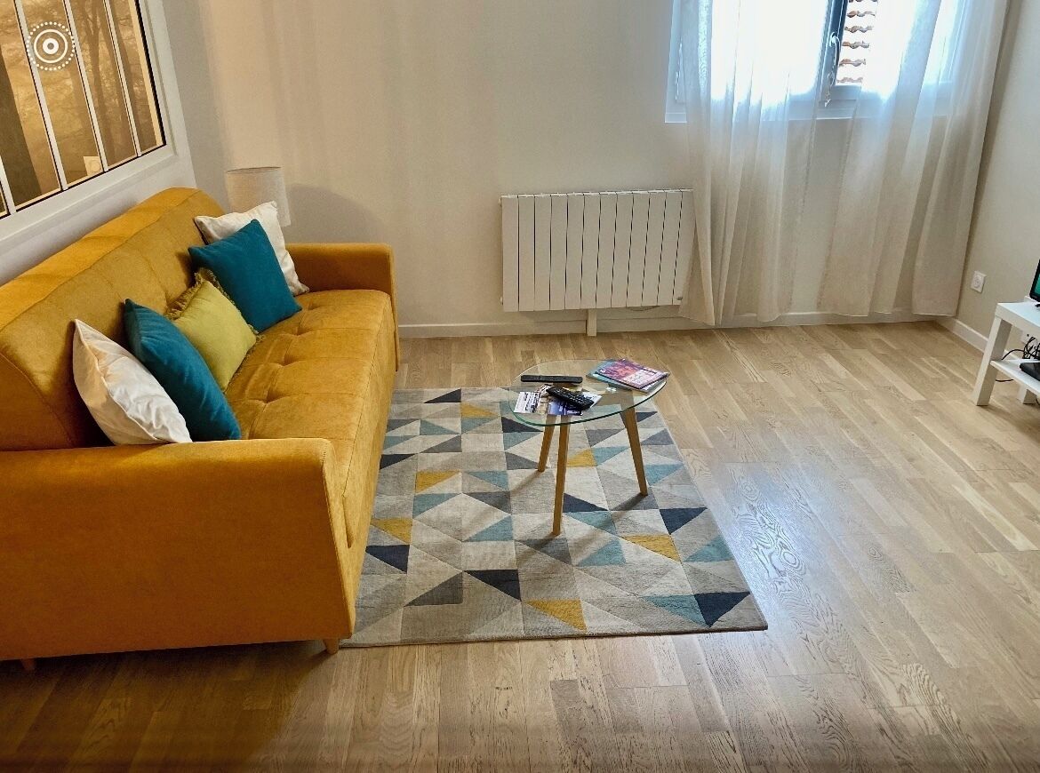 Een woonkamer met een gele bank, een vloerkleed met patroon en een kleine bijzettafel.