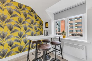 Une cuisine moderne avec un plan de travail blanc, trois tabourets, une fenêtre donnant sur des immeubles et un mur décoré d’un motif de feuilles jaune et gris.