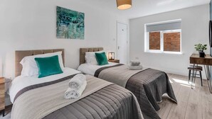 A modern hotel room with two beds, a large abstract painting, a window with brick wall view, and a small desk with a plant.