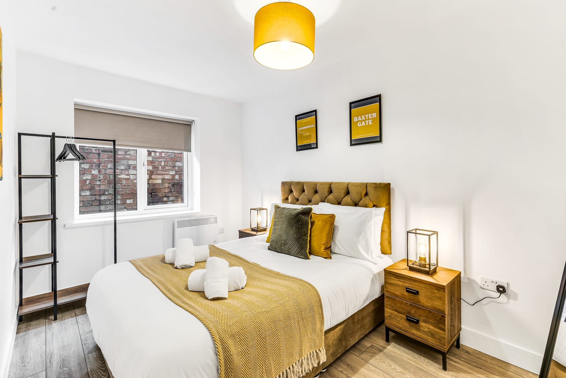 A hotel room with a bed, a bedside table, a lamp, a window with a view of a brick wall, and two framed pictures on the wall.