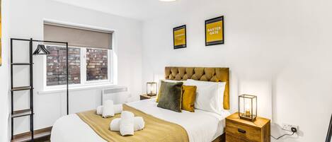 A hotel room with a bed, a bedside table, a lamp, a window with a view of a brick wall, and two framed pictures on the wall.