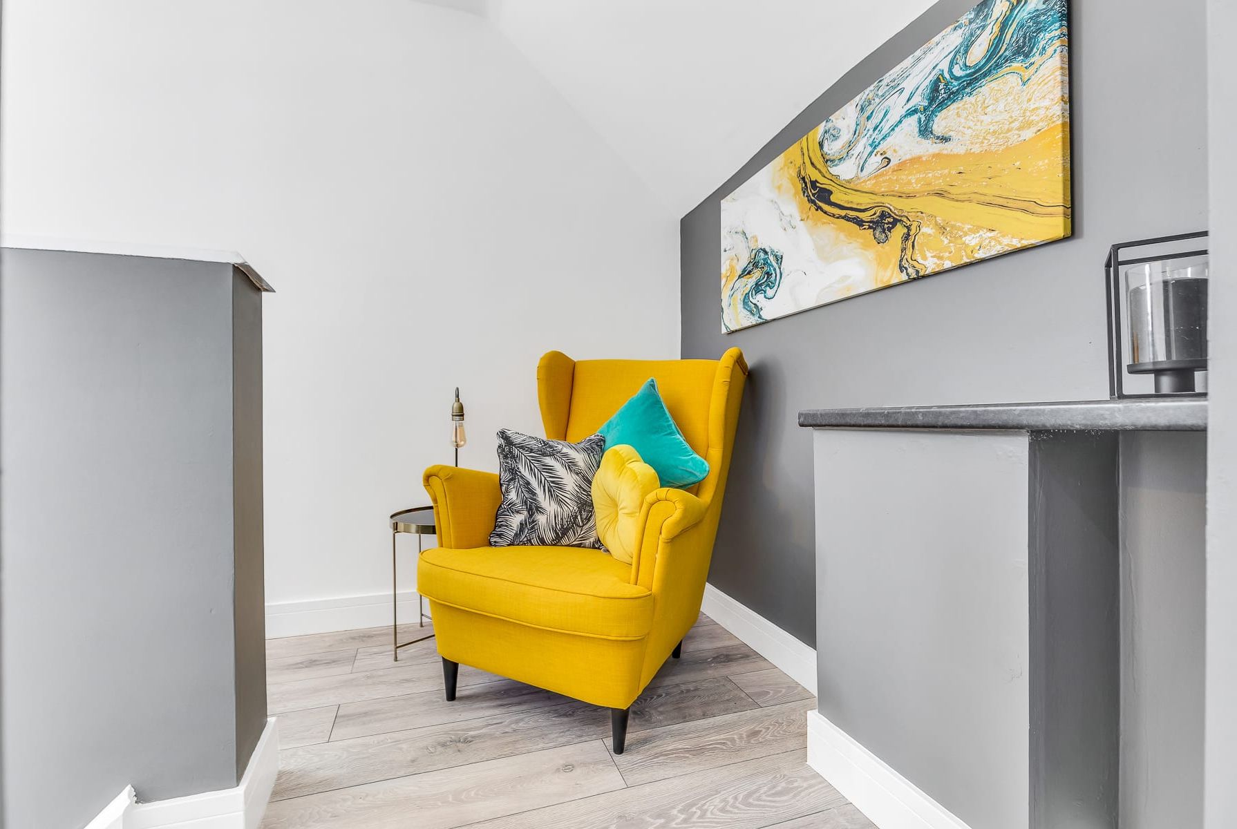 A bright yellow armchair with patterned cushions in a modern room with a grey accent wall and a piece of abstract art above it.