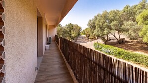 A balcony with a wooden floor, a textured wall, and a view of a landscaped area with trees and a pathway. - Apartment 4P 97M2 Sea view city center (Porto-Vecchio)