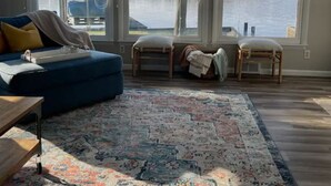 A living room with a blue sofa, two chairs, a patterned rug, and large windows overlooking a body of water. - Lakefront views at our Long Island Lookout! (Belle Center)