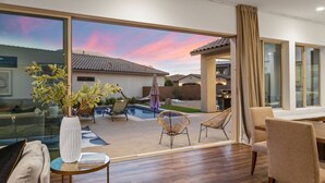 Un salon moderne doté d’une grande porte-fenêtre coulissante qui donne sur un jardin avec une piscine, du mobilier d’extérieur et une vue sur un quartier résidentiel au coucher du soleil.